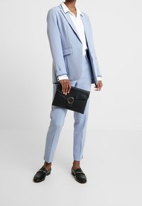 Persona che indossa un completo azzurro chiaro con camicia bianca, tiene in mano una pochette nera e indossa dei mocassini neri su uno sfondo semplice.