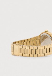 Złota metalowa bransoleta zegarka z prostokątnymi ogniwami i zapięciem, pokazana w zakrzywionej pozycji na białym tle.