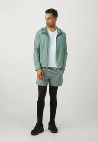 Vyras, vilkintis šviesiai žalią su kapuce striukę, baltą marškinėlį, pilkas šortus, juodas leginsus ir juodus sportinius batus, stovi prieš paprastą foną.