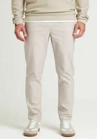 Beige maatpakken gemaakt van een gladde stof, met een slim fit en subtiele stikdetails. Gepaard met witte en grijze sneakers.