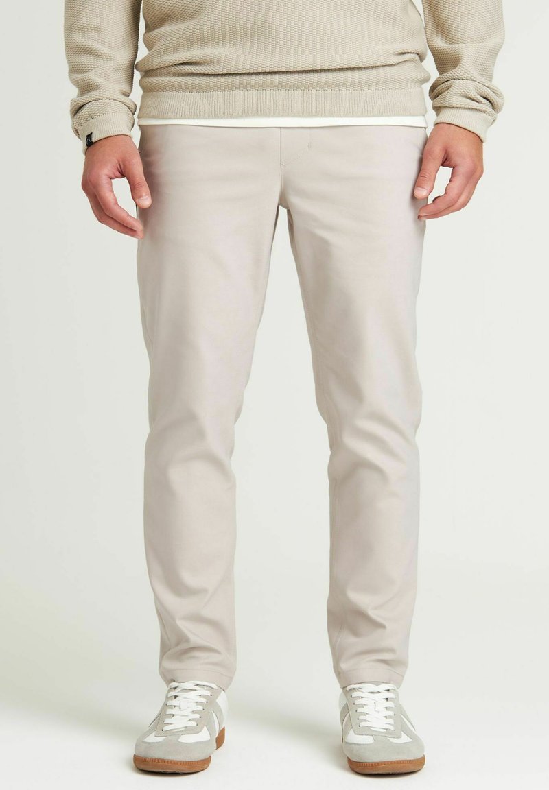 Beige maatpakken gemaakt van een gladde stof, met een slim fit en subtiele stikdetails. Gepaard met witte en grijze sneakers.