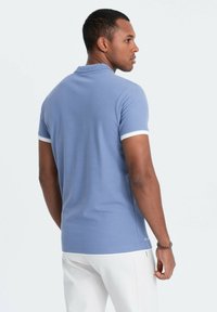 Ljusblå pikétröja med en texturerad tyg, vit krage och ärmaccents, figursydd design och diskret logotypdetalj nära fållen.