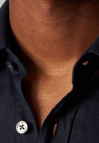 Zblízka pohled na krk a horní část hrudníku osoby, která má na sobě částečně rozepnutou tmavě modrou košili s viditelnými knoflíky.