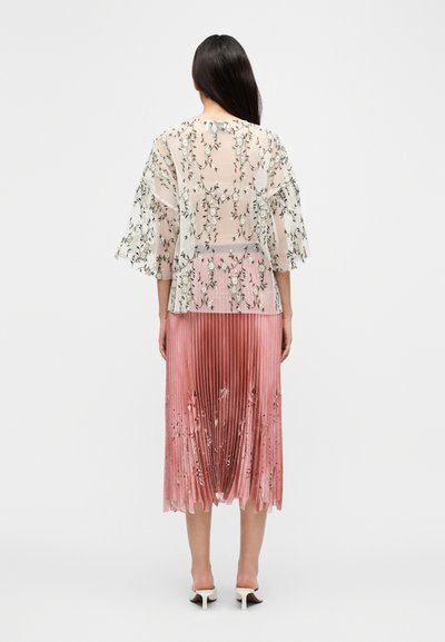 Haut transparent brodé de fleurs avec des manches évasées, associé à une jupe plissée rose ornée de motifs floraux sur la partie inférieure.