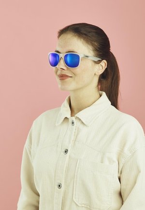 Blautönige Sonnenbrille mit einem hellgrauen Rahmen in rechteckiger Form. Die Person trägt ein beiges Cordhemd mit Kragen.