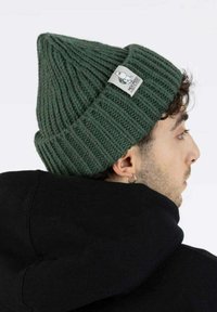 Mies, joka käyttää tummanvihreää ribattuimaista pipolakkia, jossa on valkoinen logo-tunnus, mustaa hupparia ja hopeista rengaskorua, seisoo sivuttain valkoista taustaa vasten.