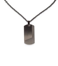 Šarnyrinė šuns ženklo pakabukas iš tamsaus metalo su įstrižomis tekstūruotomis juostomis apačioje ir dešinėje pusėje, pakabintas ant derančio grandinėlės karoliai.