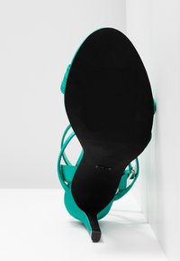 Sandale à talon haut couleur sarcelle avec une semelle noire texturée et des lanières fines pour un design ouvert. Le talon s'effile vers un bout pointu.