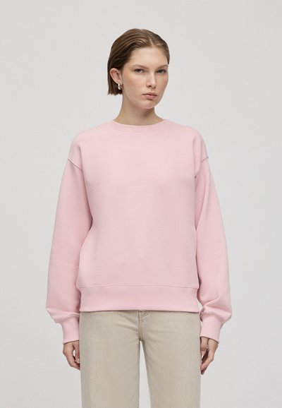 Růžová mikina s kulatým výstřihem, oversized střih a žebrované manžety. Nosené se světle béžovými kalhotami. Hladká textura, minimalistický design.