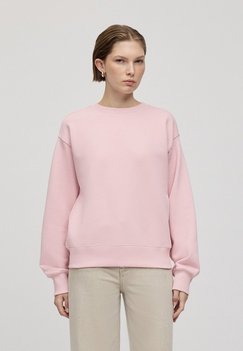 Felpa rosa con collo rotondo, vestibilità oversize e polsini a coste. Indossata con pantaloni beige chiaro. Tessuto morbido, design minimalista.
