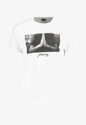 T-shirt en coton blanc avec un motif en noir et blanc représentant des mains en prière, avec "prie" imprimé en dessous dans une police cursive. Manches courtes.