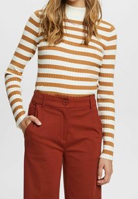 Femme portant un haut blanc et marron à rayures côtelées, manches longues, rentré dans un pantalon large taille haute couleur rouille.