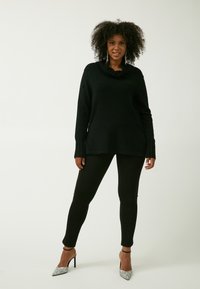 Schwarzer Strickpullover mit Rüschensaumen, langen Ärmeln und seitlichen Schlitzen. Getragen mit schwarzen Leggings und silbernen High-Heels.