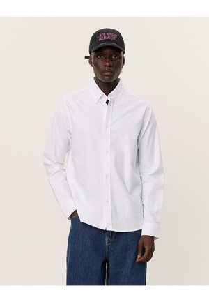 Chemise blanche à manches longues avec fermeture boutonnée, col pointu et manches à poignet, assortie à un pantalon en denim bleu et une casquette noire avec du texte.
