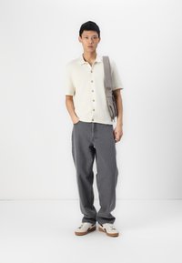 Chemise crème à manches courtes avec des boutons, jean oversized gris et baskets blanches avec des accents marron. Portant un sac à bandoulière gris.
