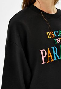 Svart sweatshirt med färgglad broderad text som säger "FLY IN I PARADIS" på bröstet, bärs av en person med brunt hår.