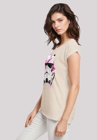 Beige T-shirt with a graphic print of a Stormtrooper helmet in black, white, and pink. Short sleeves and relaxed fit, made of soft fabric.