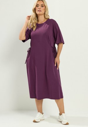 Femme aux longs cheveux blonds portant une robe midi violette ample à manches courtes et des baskets blanches, debout devant un fond uni.