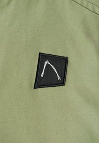 Olijfgroen stof heeft een zwart vierkant patch met witte lijntekeningen. Het patch is stevig gestikt en toont een minimalistisch ontwerp.