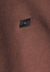 Tejido marrón texturizado con un logotipo bordado negro que asemeja un rectángulo, colocado en el centro con un diseño limpio.