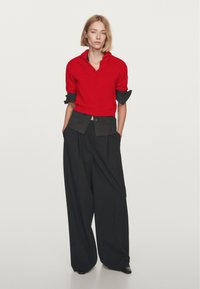 Donna con capelli biondi che indossa un maglione rosso brillante a maniche corte sopra una camicia nera a maniche lunghe e pantaloni neri larghi, con le mani in tasca, su uno sfondo neutro.