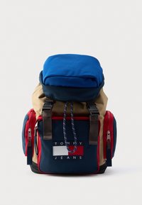 Rucsac cu clapetă superioară albastră, corp kaki și accente în albastru închis și roșu. Are un patch cu logo și mai multe compartimente cu fermoar.