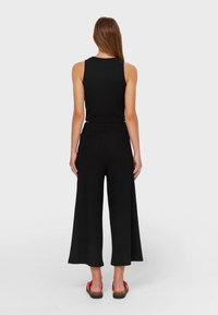 Kvinna som står med ryggen mot kameran iförd en svart ärmlös topp, vida svarta byxor och röda sandaler på en neutral bakgrund.