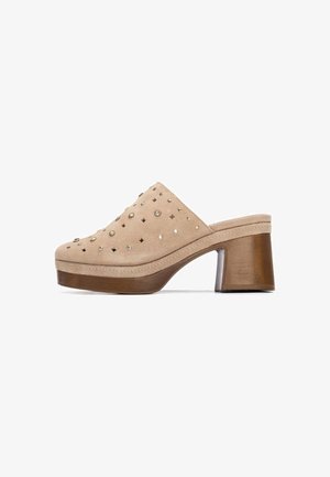 Zueco de ante beige con puntera puntiaguda, adornado con tachuelas doradas y plateadas, tacón de bloque de madera y laterales suaves.