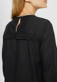 Schwarze Bluse aus einem strukturierten Stoff mit vertikalen Streifen, einem Schlüsselaugen-Rücken und einer gewellten Spitze entlang der Schultern.