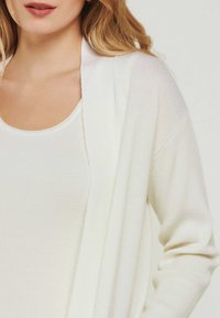Weiße offene Strickjacke mit langen Ärmeln und gerippter Textur, über einem weißen, eng anliegenden Tanktop getragen. Weiches, leichtes Material.