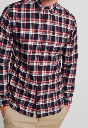 Homme portant une chemise à carreaux bleu marine, rouge et blanc boutonnée, avec un pantalon beige, mains dans les poches, sur fond gris clair.