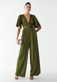 Ženska stoji v širokem olivno zelenem kombinezonu z globokim V-izrezom in plavajočimi rokavi, drži majhno zlatih ročno torbico in nosi zlate uhane v obliki obroča.