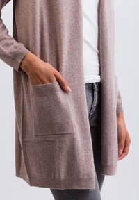 Lichtbruine cardigan met lange mouwen, geribbelde manchetten, open voorkant en een voorkeepocket. Zachte textuur en een ontspannen pasvorm. Gedragen over een witte top.