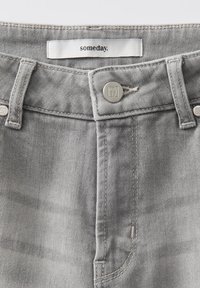 Close-up van de lichtgrijze spijkerbroek tailleband met metalen knoop en een wit label met de tekst "someday" boven de knoop.