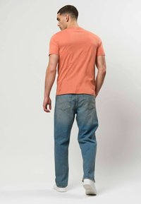 Homme vu de dos portant un t-shirt manches courtes couleur saumon, un jean bleu et des baskets blanches sur un fond clair uni.