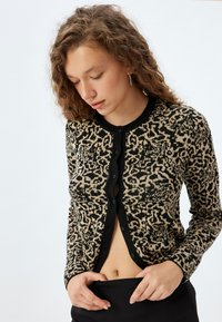 Cardigan con stampa leopardata nera e beige, dotato di una chiusura con bottoni, colletto a costine e maniche lunghe. Tessuto a maglia testurizzato e design aderente.