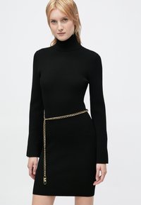Fekete bordázott ruha hosszú ujjal és magas nyakkivágással, arany lánc övvel kiemelve. Az anyag texturált felülettel rendelkezik.