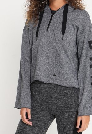 Vrouw draagt een grijze hoodie met lange mouwen en zwarte trekkoorden, samen met donkergrijze nauwsluitende leggings, staand met ontspannen handen langs haar zij.