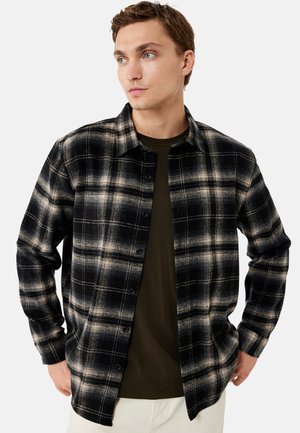 Giovane uomo che indossa una camicia a quadri nera e beige abbottonata sopra una t-shirt verde oliva, in piedi con le mani nelle tasche, guardando di lato.