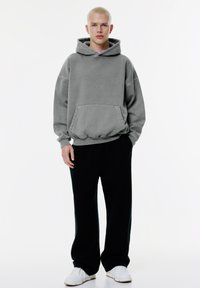 Felpa grigia oversize con tasca frontale e cappuccio regolabile con cordino, abbinata a pantaloni neri larghi e sneakers bianche.