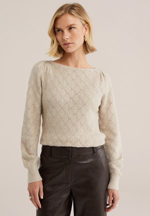 Femme blonde portant un pull en maille beige texturé et un pantalon en cuir marron foncé, regardant légèrement sur le côté sur un fond uni.