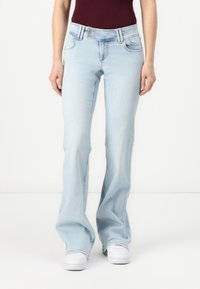 Jeans evazate de culoare bleu deschis, cu un finisaj decolorat, două buzunare frontale și închidere cu fermoar, asortați cu adidași albi.