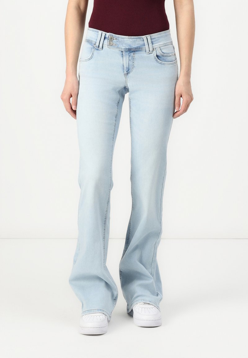 Jeans evazate de culoare bleu deschis, cu un finisaj decolorat, două buzunare frontale și închidere cu fermoar, asortați cu adidași albi.