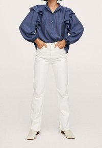 Blue button-up shirt with ruffled sleeves, paired with white straight-leg jeans and beige ankle boots. Smooth fabric, relaxed fit.