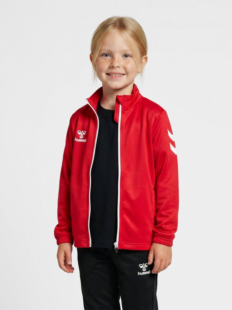 Joven sonriente con cabello rubio que lleva una chaqueta deportiva roja con cremallera y pantalones negros con logotipos blancos de hummel sobre un fondo liso.