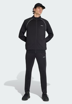 Mann trägt schwarzen Adidas-Trainingsanzug und Kappe, steht mit den Händen hinter dem Rücken auf hellem Hintergrund.