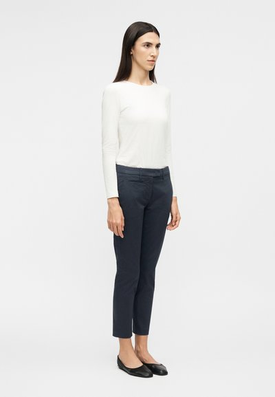 Γυναίκα που στέκεται με κατεύθυνση ελαφρώς προς τα δεξιά, φοράει άσπρο μακρυμάνικο τοπ, σκούρο στενό παντελόνι μήκους μέχρι τον αστράγαλο και μαύρα επίπεδα παπούτσια, σε λευκό φόντο.