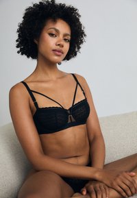Black lace bra featuring scalloped edges, decorative straps, and gold accents. Soft texture with a structured design.