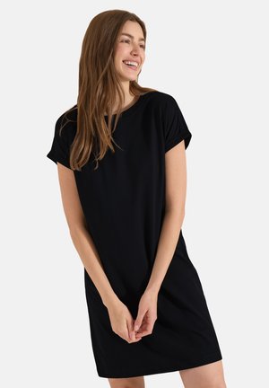 Junge Frau lächelnd, trägt ein einfaches schwarzes Kurzarmkleid, steht mit gefalteten Händen vor einem schlichten hellen Hintergrund.