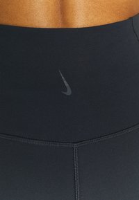 Schwarze, hochtaillierte Leggings aus elastischem Material. Mit einem subtilen dunkelgrauen Nike-Logo. Glatte Textur mit kontrastierenden Nähten.